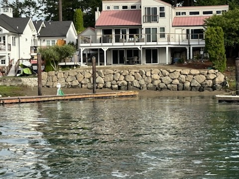 Modern waterfront home with red metal roof, large boulder retaining wall, and dock with buoy in the Puget Sound region