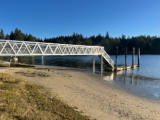 White metal pedestrian bridge extending to a floating dock on calm water, surrounded by evergreen forest in the Salish Sea region